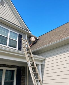 IMG_3223 | Wildlife Control Specialist of Atlanta Worker on a tall ladder performing roof work along the eaves of a beige house under a clear blue sky.