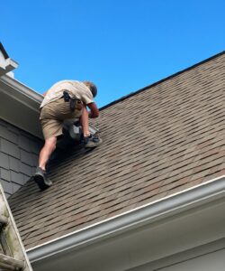 IMG_3224 | Wildlife Control Specialist of Atlanta Worker on a sloped shingle roof crouching near a white gutter, installing or inspecting it under a clear blue sky.